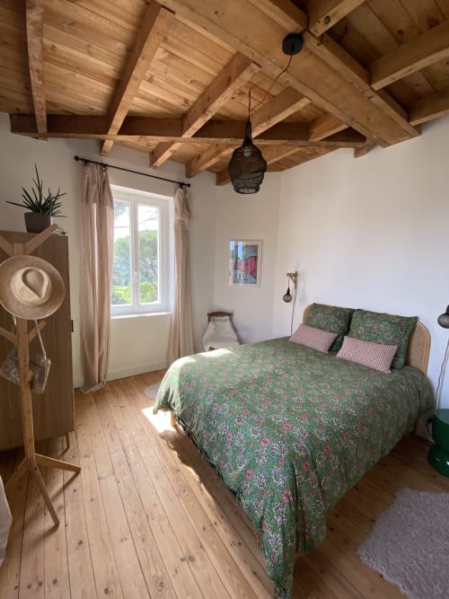 Chambre rustique au Domaine les Olivoux à Pomport avec poutres apparentes, literie fleurie verte et grande fenêtre.