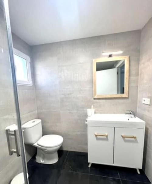 Baño moderno en Apartamento Jacaranda de ROILA360 Alicante con lavabo blanco, espejo, inodoro y suelo de baldosas negras