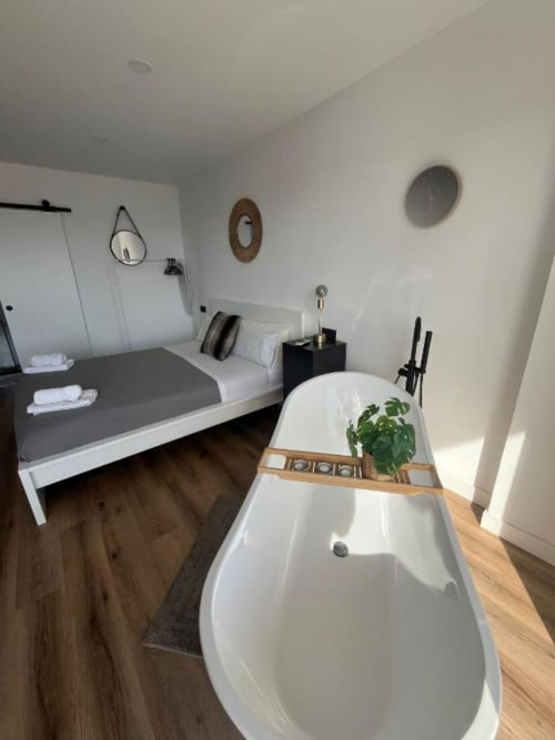 Dormitorio moderno con bañera exenta, cama blanca, espejos y suelos de madera oscura en apartamento de Alicante