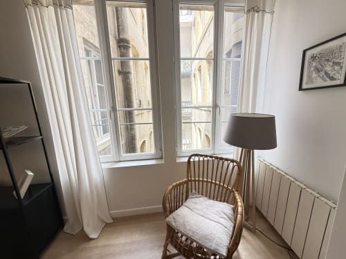 Chambre lumineuse d'un appartement à Metz avec fauteuil en rotin, lampadaire et vue sur la ville par de grandes fenêtres aux voilages blancs