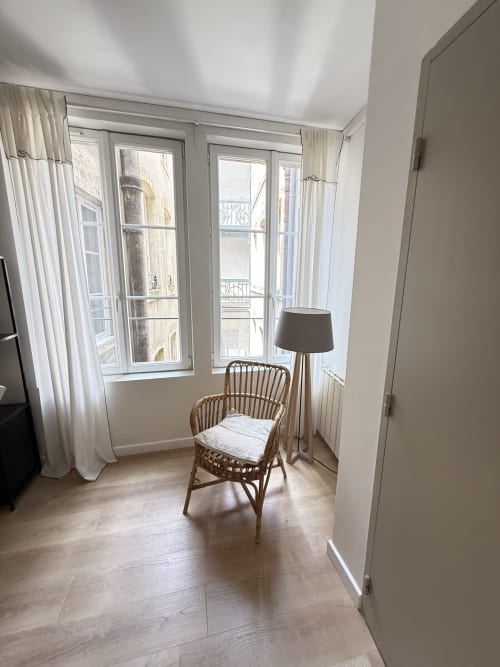 Chambre lumineuse chez Mattbnb Metz avec fauteuil en rotin, lampadaire et vue sur la ville par les fenêtres