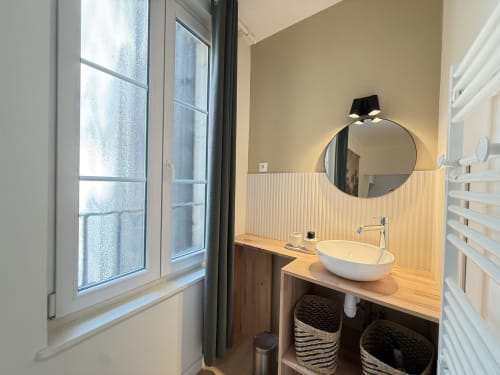 Salle de bain moderne dans un appartement de Metz avec miroir rond, lavabo blanc, plan de travail en bois et vue sur la fenêtre