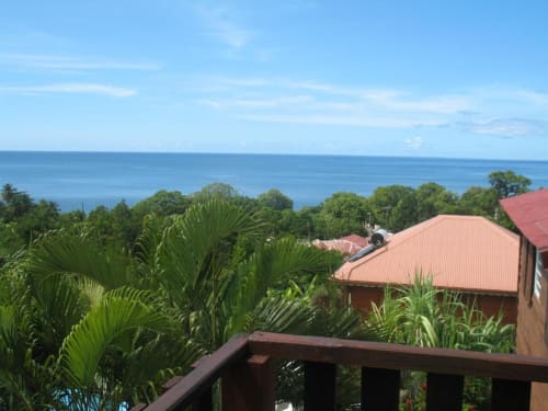 Vue depuis la terrasse du Bungalow Kénet sur l'océan, jardins tropicaux et bâtiments aux toits rouges d'Art Lodge Kafé, Bouillante