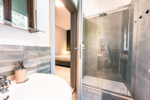 Bagno moderno con doccia aperta presso Casa Giulietta, Como. Box doccia in vetro, piastrelle grigie, lavabo bianco, vista sulla camera da letto