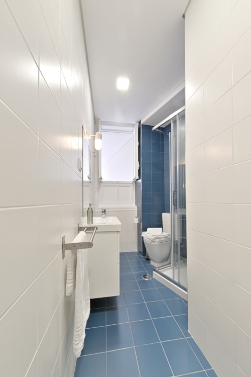 Modern bathroom with white wall tiles, blue floor tiles, sink, toilet, shower enclosure, and window in Porto apartment