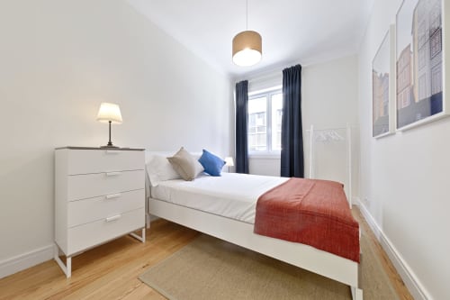 Modern bedroom in downtown Porto apartment with white bed, blue curtains, red throw blanket, wooden dresser, and city views