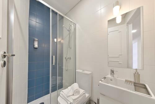 Modern bathroom in downtown Porto apartment with glass shower enclosure, white tiles, blue accent wall, and illuminated