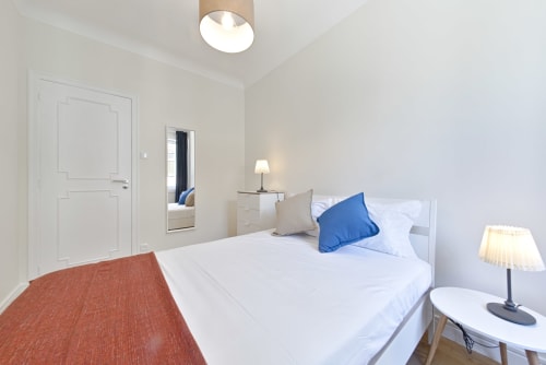 Quarto moderno em apartamento do Porto com cama branca, almofada azul, luminária de cabeceira, espelho e tapete terracota no Aveiro com Paixão