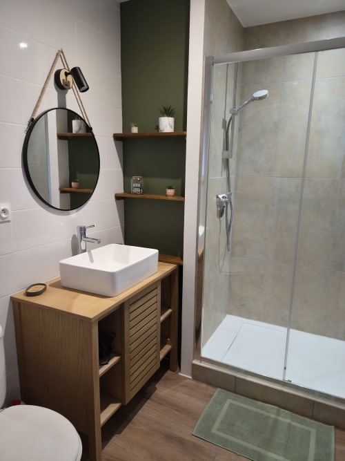 Baño moderno con lavabo blanco de cerámica, tocador de madera, espejo redondo, pared acentuada en verde oliva y ducha tipo walk-in en Domaine