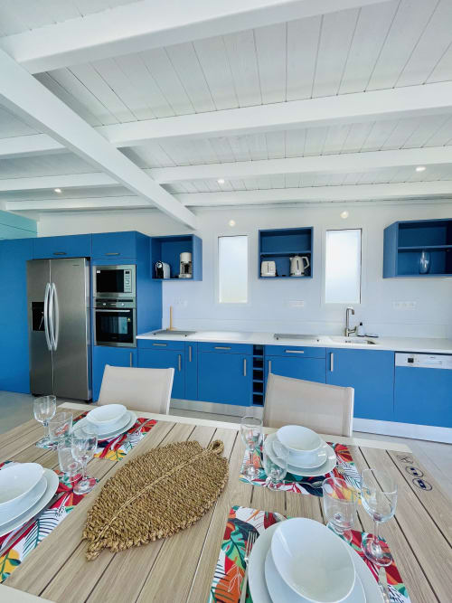 Cuisine moderne bleue avec plafond à poutres blanches, table à manger dressée avec sets de table colorés à la Villa Nid des