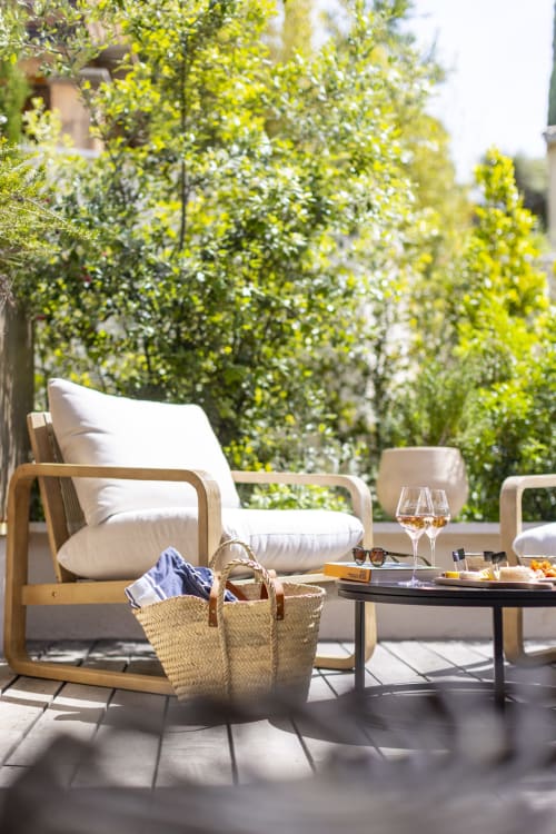 Terrasse ensoleillée de La Maison des Jardins avec chaise longue en bois, panier tressé et verres de vin face au lierre verdoyant