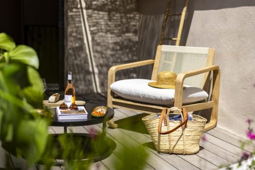 Terrasse ensoleillée de La Maison des Jardins avec chaise longue en bois, paniers de paille, bouteille de vin et plantes fleuries