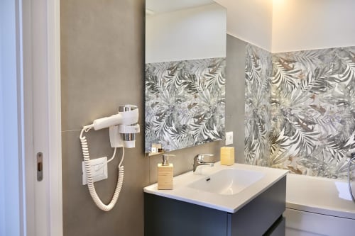 Bagno moderno a Taga Rooms Catania con piastrelle decorate, vanity scuro, lavabo bianco, specchio e asciugacapelli a parete