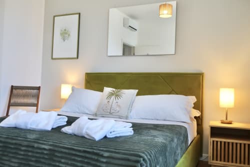 Camera moderna di Taga Rooms Catania con letto imbottito verde oliva, cuscini bianchi, lampade laterali, specchio e vista sulla città