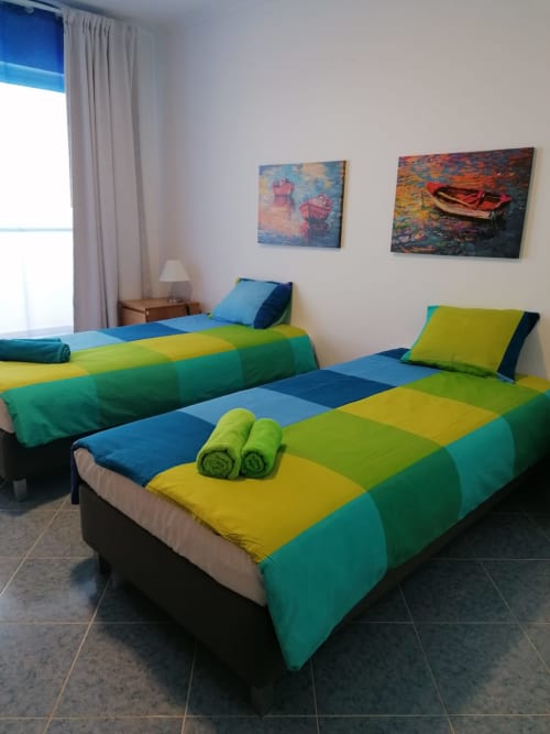 Quarto iluminado com duas camas com roupa de cama em azul, verde e amarelo, toalhas dobradas e quadros de barcos em A Casa