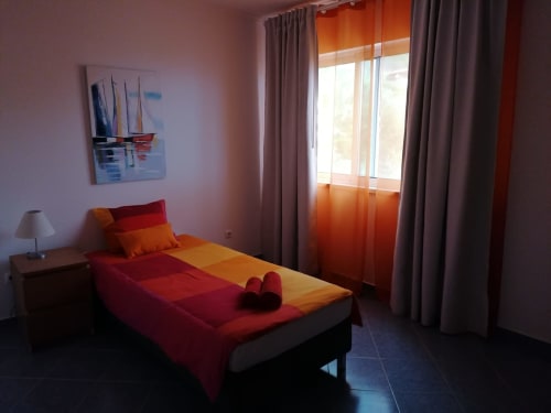 Quarto luminoso em A Casa da Xã com roupa de cama vermelha e laranja, janela com vista para o mar com cortinas laranja translúcidas, quadro de veleiro e