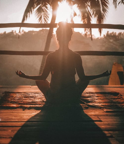 Femme en méditation au coucher de soleil sur une terrasse en bois avec palmiers, refuge tropical Bleu Guesthouse