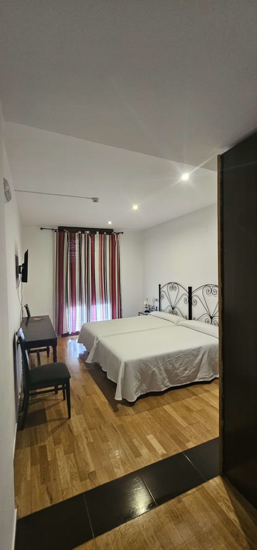 Luminosa habitación doble en Hostal El Pasaje con dos camas, cortinas burdeos, piso de madera, TV y moderna iluminación