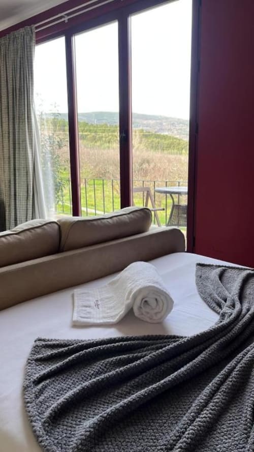 Quarto com sofá cinzento, toalhas enroladas na cama e vista para a serra através das portas de vidro da pousada Tranca da Barriga, Covilhã