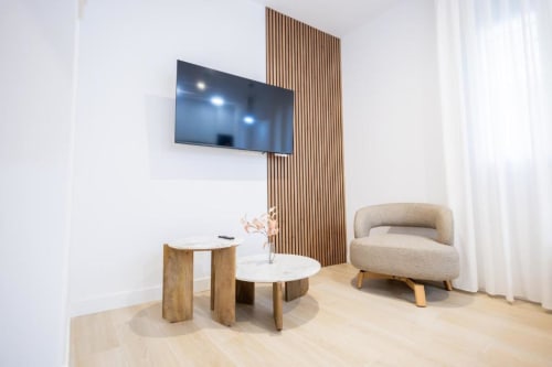 Moderna sala de estar en apartamento La Búsqueda, Cuenca con TV de pared, muro de madera, butaca beige y mesas laterales