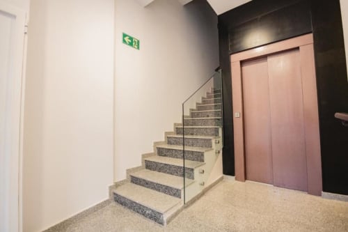 Entrada de moderno edificio residencial en Cuenca con escaleras, barandilla de cristal, puertas de ascensor rosa y salida de emergencia verde