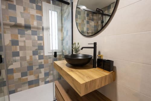 Baño moderno con lavabo negro de cerámica, vanidor de madera, grifo negro, espejo redondo y ducha con azulejos geométricos en Cuenca