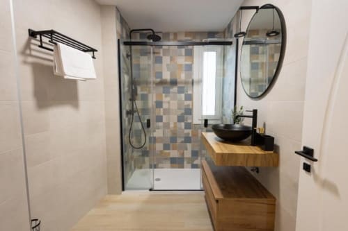 Baño moderno en apartamento de Cuenca con ducha abierta, accesorios negros, tocador de madera y espejo redondo