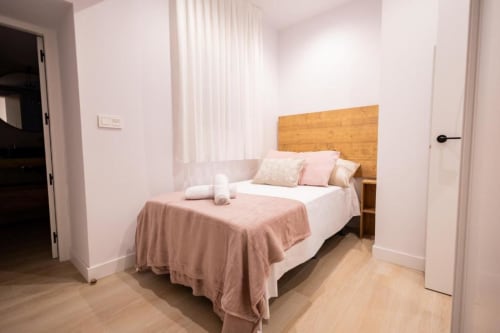 Dormitorio luminoso en apartamento de Cuenca con cabecero de madera, ropa de cama blanca, detalles en rosa y piso de madera clara