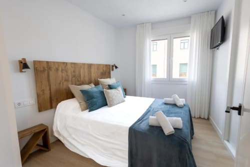 Dormitorio moderno en apartamento de Cuenca con cabecero de madera, acentos azules, ropa de cama blanca, TV de pantalla plana y vistas a la ciudad