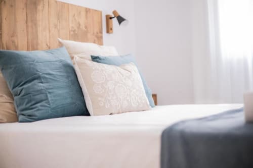 Dormitorio moderno con cabecera de madera, cojines decorativos azules y crema, ropa de cama blanca y lámpara de pared en Cuenca
