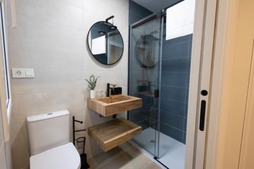 Baño moderno en Apartamentos La Búsqueda, Cuenca. Lavabo de madera, espejo redondo, ducha alicatada, inodoro y planta verde.