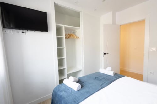 Dormitorio moderno en apartamento de Cuenca con cama azul, ropa de cama blanca, televisor de pantalla plana, armario y puerta de madera