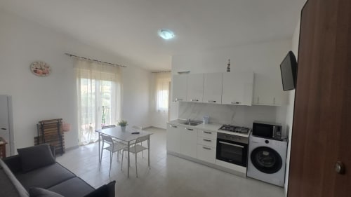 Cucina e zona living open-plan in moderno appartamento, con mobili bianchi, tavolo da pranzo e accesso al balcone con vista mare a Tronca