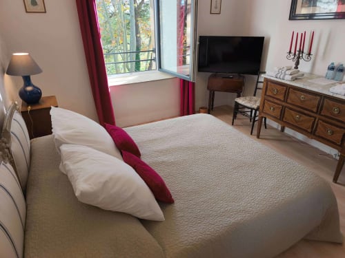 Elegant bedroom at Magnanerie de Sainte Croix with garden views, white bed, red accents, TV, wooden dresser, and bright
