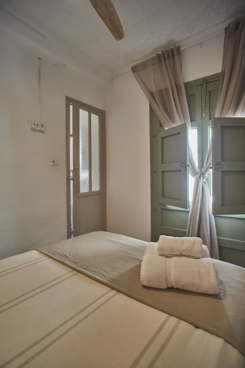Habitación luminosa en Casas Cercana con cama doble, toallas enrolladas, puertas de madera, persianas verdes con cortinas y ventilador de techo.