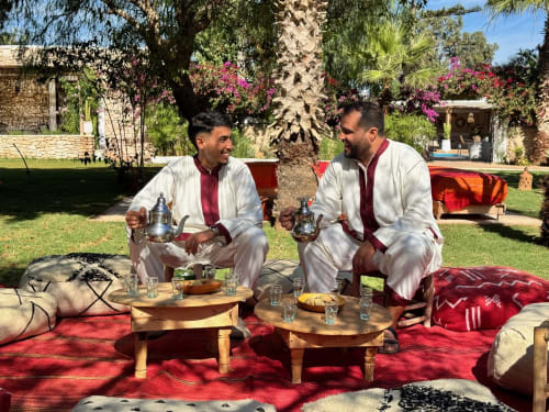 Deux hommes en tenue marocaine traditionnelle servant le thé au Riad Eucalyptus à El Ghazouane, détendus sur des coussins rouges dans un jardin