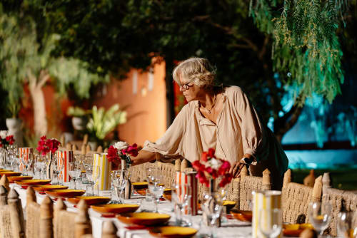 Femme dressant une table de repas en plein air au Riad Eucalyptus avec fleurs rouges, verres et décoration traditionnelle à El Ghazouane