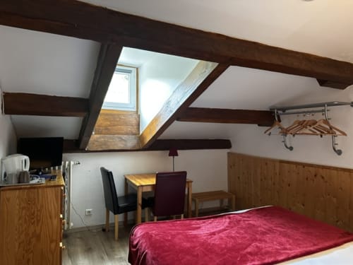 Chambre mansardée à l'Hôtel la Source avec poutres apparentes, fenêtres de toit, lit aux draps rouges, bureau et mobilier en bois