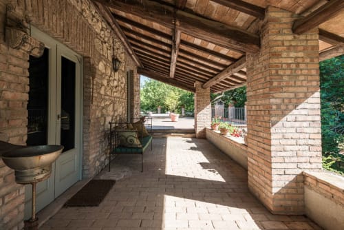 Patio coperto con muri in laterizio rosso e travi in legno presso l'appartamento Il Casale a Viterbo, con sedute esterne e giardino