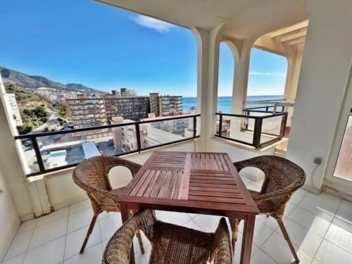 Terraza soleada con mesa de comedor de madera y sillas de mimbre con vistas al mar Mediterráneo en apartamento de Fuengirola