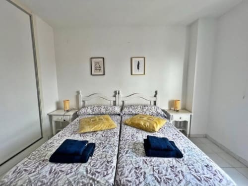 Dormitorio moderno y luminoso en apartamento de Fuengirola con dos camas gemelas, almohadas amarillas, toallas azules, mesillas blancas y cuadros