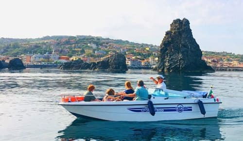 Turisti su una motobarca che esplorano la drammatica costa rocciosa di Aci Castello con edifici colorati e faraglioni.
