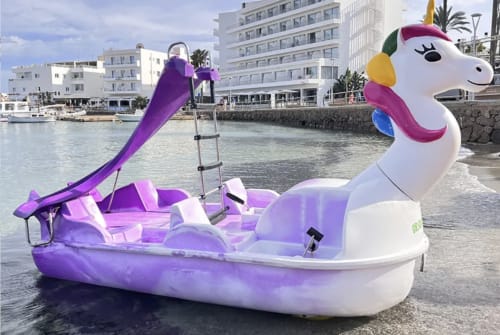 Bote de pedales en forma de unicornio morado y blanco en la playa del hotel Playa Sol en Es Canar, Ibiza, con edificios costeros