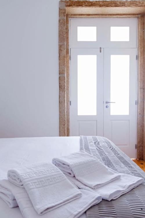 Toalhas e roupas de cama brancas cuidadosamente dobradas na cama do Bom Porto Apartments, com portas de vidro e moldura de madeira no Porto.