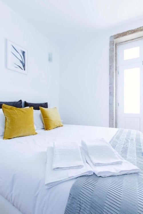 Quarto luminoso no Bom Porto Apartments com cama branca, almofadas amarelas e porta com vista para o jardim no apartamento do Porto.