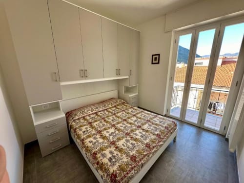 Camera moderna con armadio integrato, letto con biancheria a fantasia e balcone con vista sui tetti di Finale Ligure