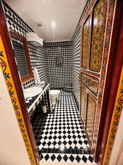 Salle de bains marocaine du Riad Dar Tahri à Fès avec carrelage noir et blanc, lavabo, toilettes et décoration rouge et or