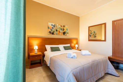 Camera moderna La Zagara Rooms, Milazzo con letto in legno, biancheria bianca, cuscini verdi, illuminazione calda e vista città