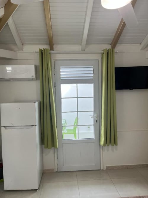 Intérieur lumineux du bungalow à la Résidence B&L Lagon avec électroménagers blancs, rideaux verts et vue jardin par porte vitrée