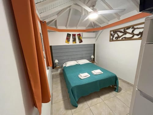 Chambre Bungalow Corail à la Résidence B&L Lagon avec lit turquoise, rideaux orangés et vue sur le jardin en Martinique